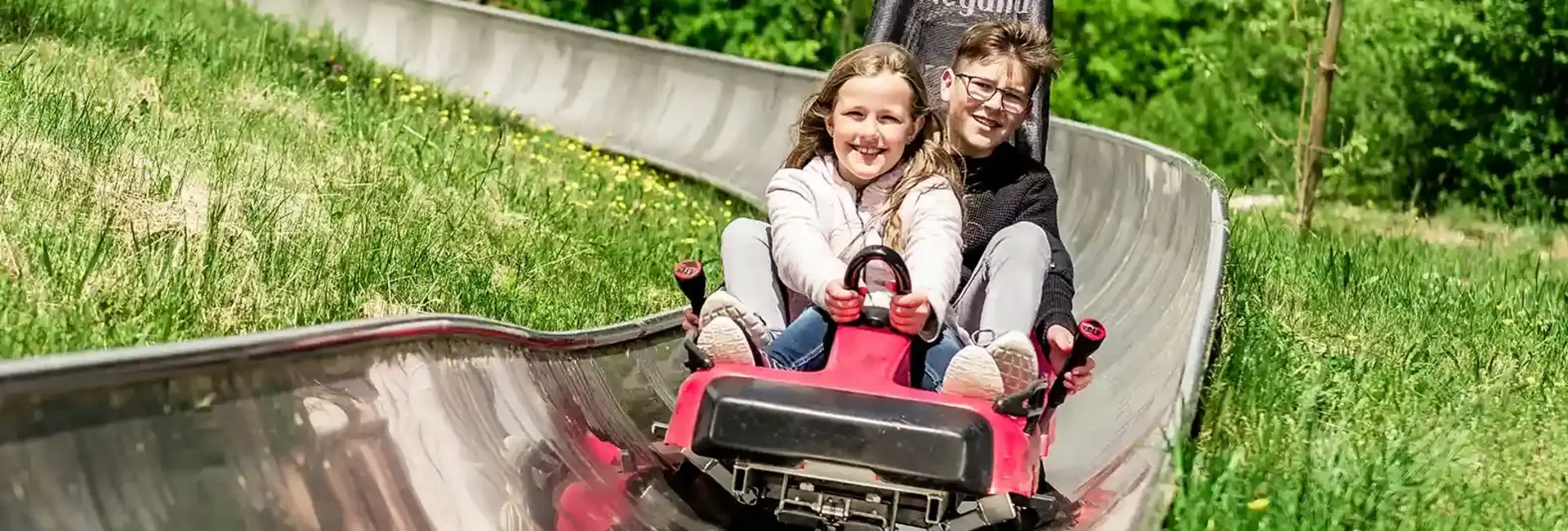 Zwei Kinder fahren mit der Sommerrodelbahn inmitten der Natur. Beide sind am grinsen und halten sich am Schlitten fest. Der Junge hat die Bremshebel in der Hand um die Geschwindigkeit selbst zu steuern.